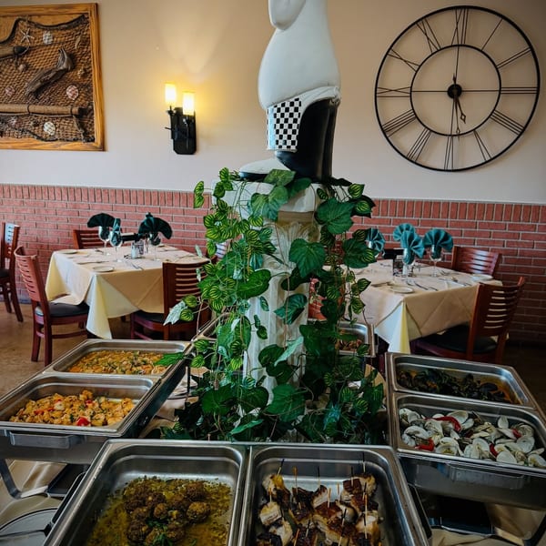 Buffet line with various dishes, set in a restaurant dining room with tables and a chef statue.