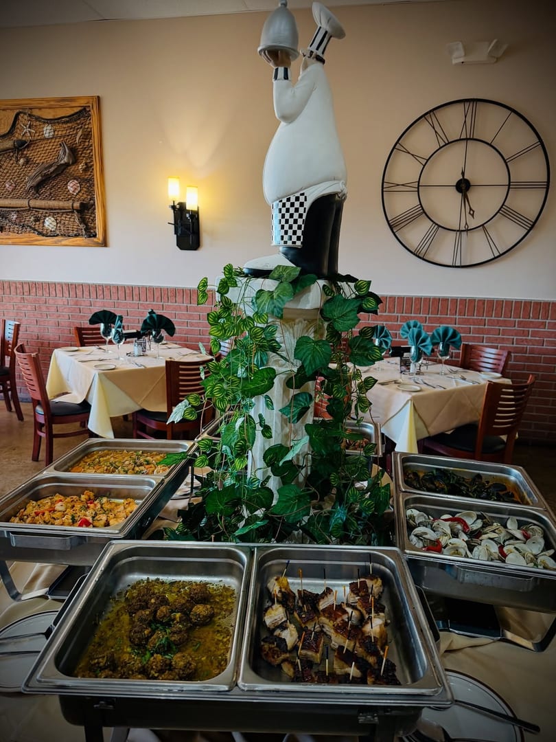 Buffet line with various dishes, set in a restaurant dining room with tables and a chef statue.